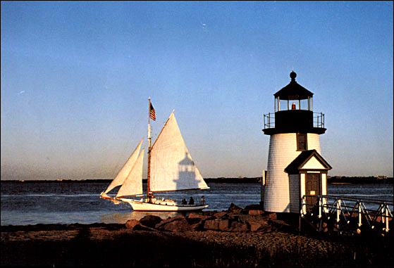 Small Gaff-rigged sailboat under mainsail and two jibs passing a white lighthouse which is making a shadow outline of itself on the sail